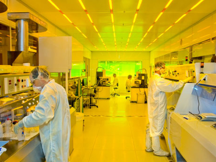 Four people in PPE working at different stations in a narrow clean room bathed in yellow light